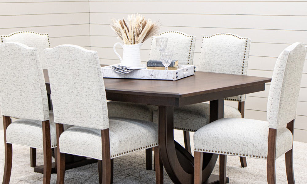 A dark wood dining table with six light upholstered chairs, nailhead trim, glassware, books, and a white vase.
