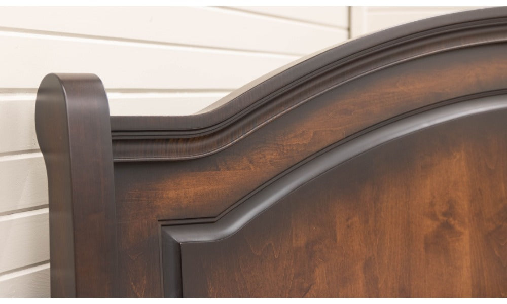 A close detail of a dark wooden bed headboard with curved molding against a light paneled wall and visible wood grain.